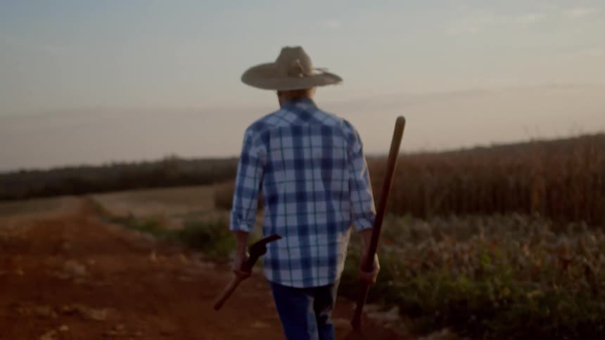 Farmer walking down a dirt path in field with tools at sunset