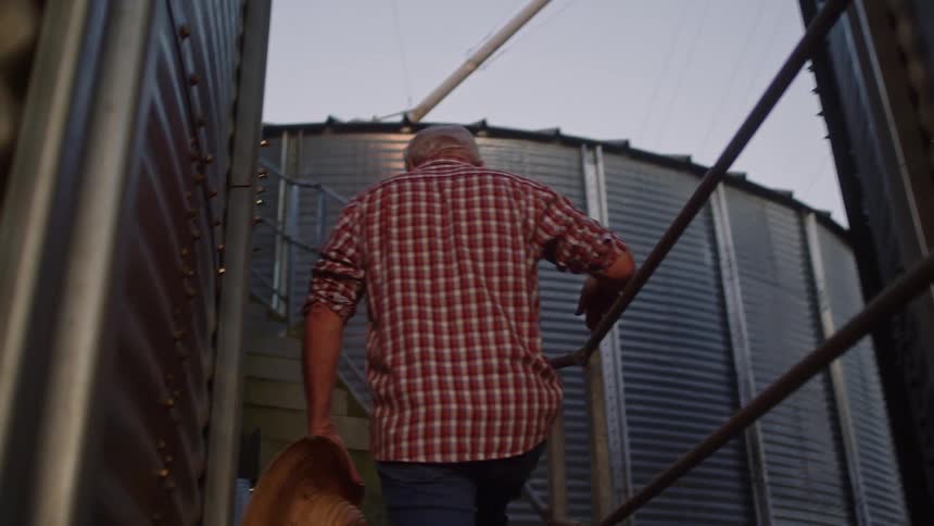A man wearing a red plaid shirt and blue jeans ascends stairs between two large metal silos holding a straw hat in his hand