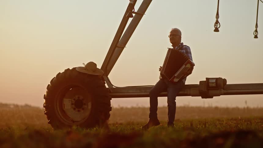 A man plays the accordion while sitting on farm irrigation equipment at sunset