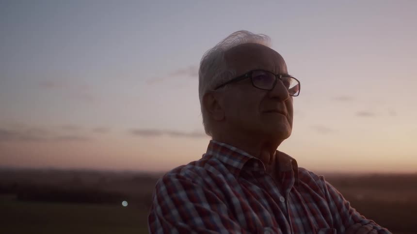 A man with grey hair and glasses stands with his arms crossed looking towards the right side of the frame with a soft sky in the background
