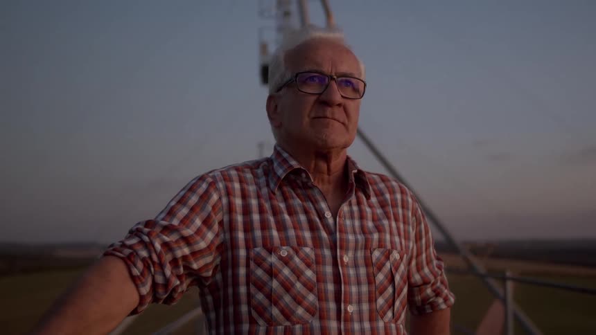 An elderly man wearing glasses stands on a pedestrian bridge at a construction site