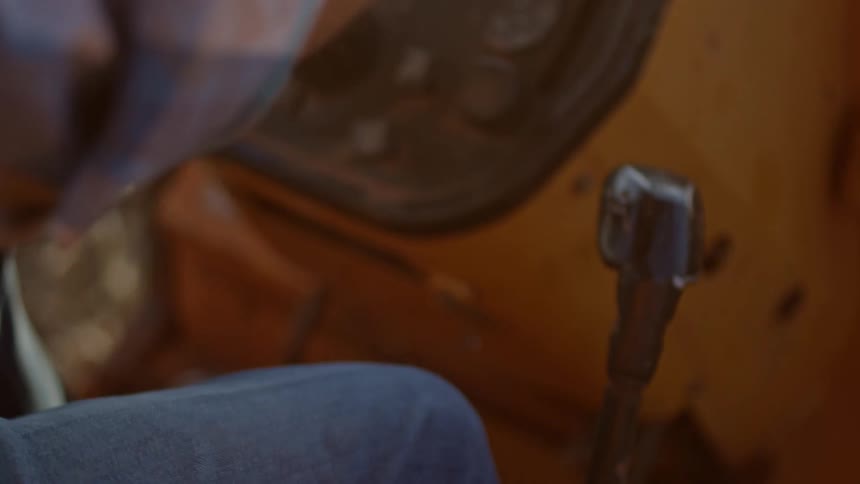 A person operates the control lever of a heavy machine or vehicle with part of the dashboard and worn interior visible in warm lighting