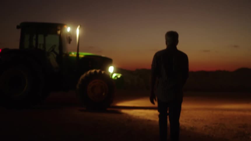 Man walks towards a farm tractor with lit headlights parked outdoors at night