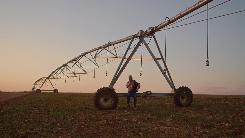A man stands on tilled soil playing an accordion beneath a long metal irrigation system with a clear sky in the background