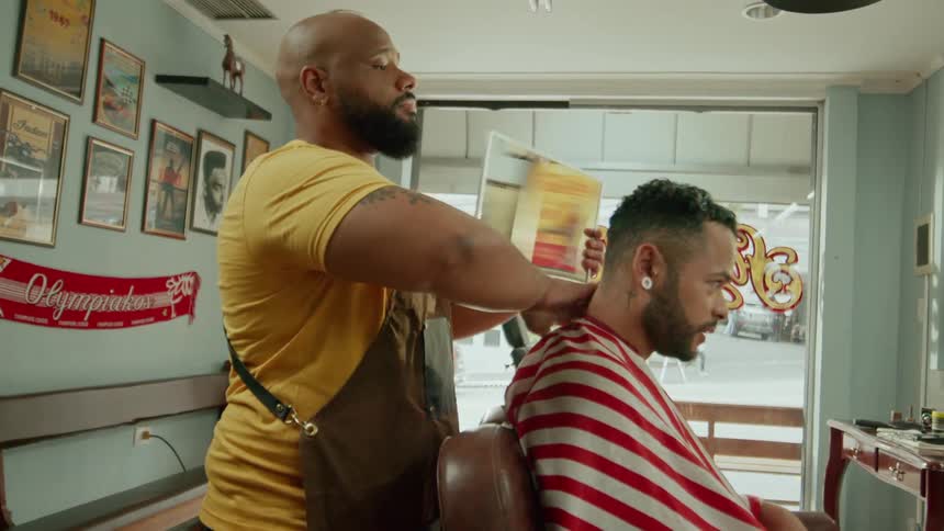 A barber wearing a yellow shirt and apron is giving a haircut to a client draped in a redandwhite striped cape inside a vintagestyle barbershop