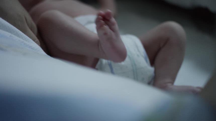 A baby wearing a diaper lifts its foot into the air as a hand smooths the bed underneath