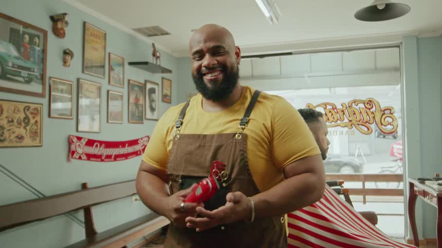 A smiling barber holds clippers with a client seated behind him in the shop