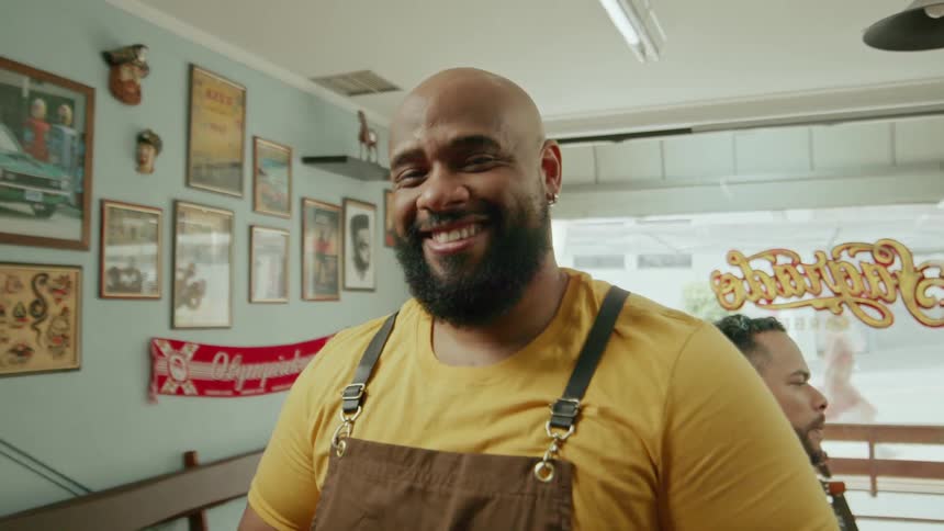 A barber holds a red clipper with a smile on his face while a customer in the barbering chair behind him checks out his haircut in the mirror at midday