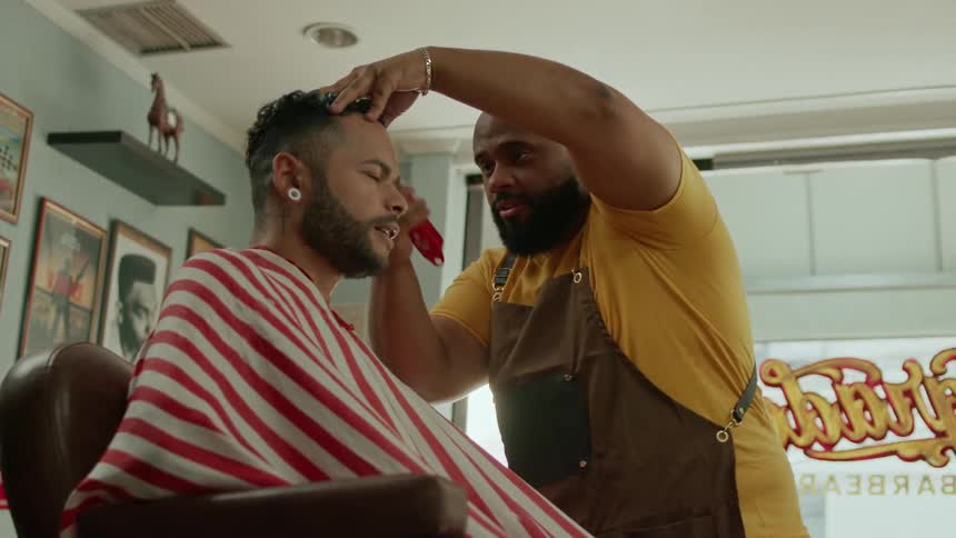 A smiling barber gives a cheerful client a haircut in a warmly lit barbershop