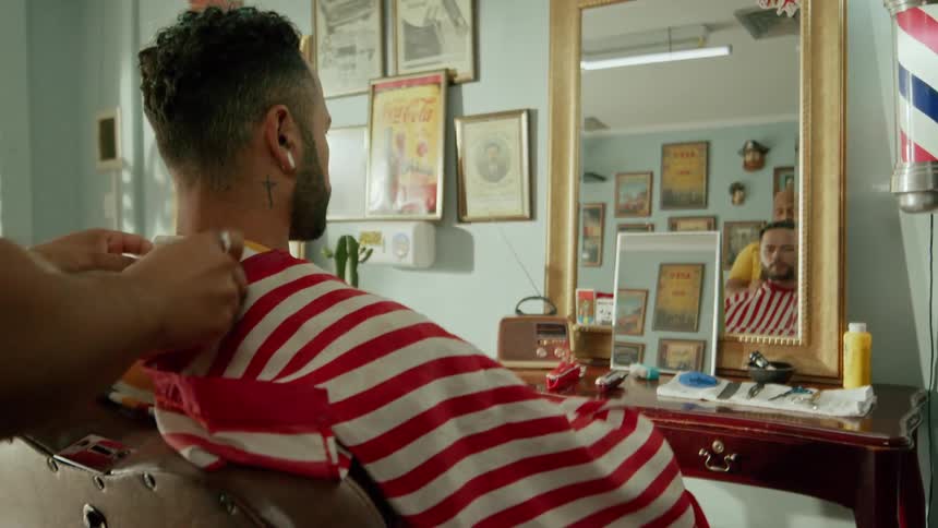 A barber drapes a red and white striped cloth around the neck of a person in a colorful striped shirt seated in a barbers chair