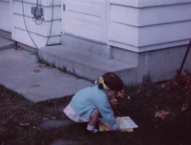 A toddler carries books around outside and sits down