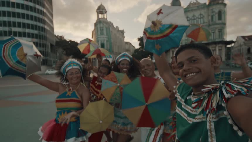 A joyful group dressed in colorful traditional costumes holds vibrant umbrellas while smiling for a selfie in a lively city square at sunset