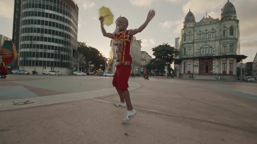 A person in vibrant clothing leaps into the air while dancing in a city square holding a yellow umbrella