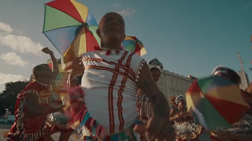 A group of people in native attire dance in the middle of the street holding small umbrellas