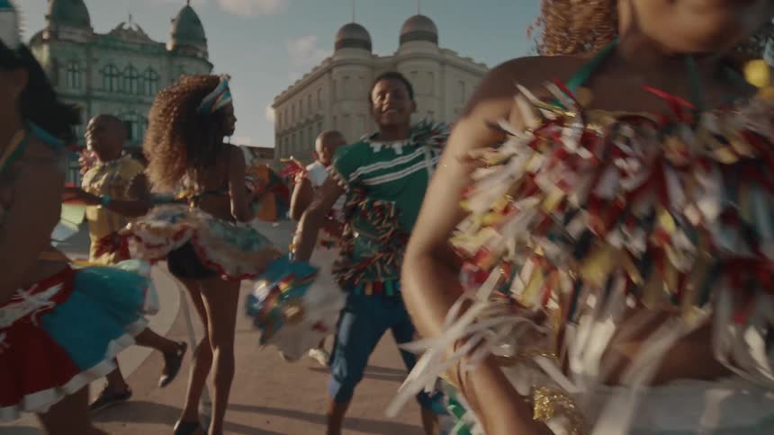A vibrant outdoor celebration with several people dancing in colorful traditional attire holding small multicolored umbrellas they are moving joyfully through what appears to be a city square with historic buildings in the background