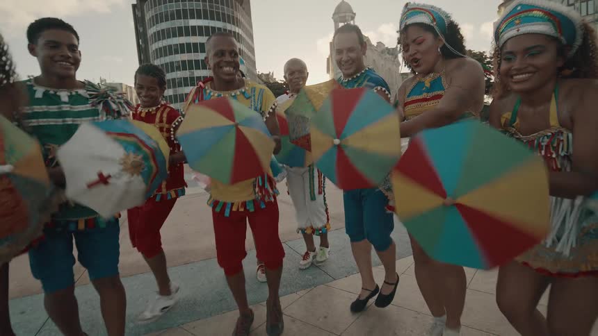 A group of people wearing vibrant colorful costumes and holding small multi colored umbrellas are dancing outdoors in a paved urban area with buildings in the background