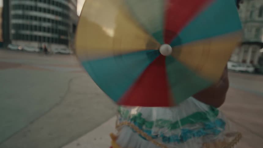 A woman dressed in carnival clothing holds a small rainbowcolored umbrella and dances joyfully by the roadside at midday