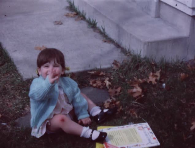 An older film of  a child sitting outside on the sidewalk and yard