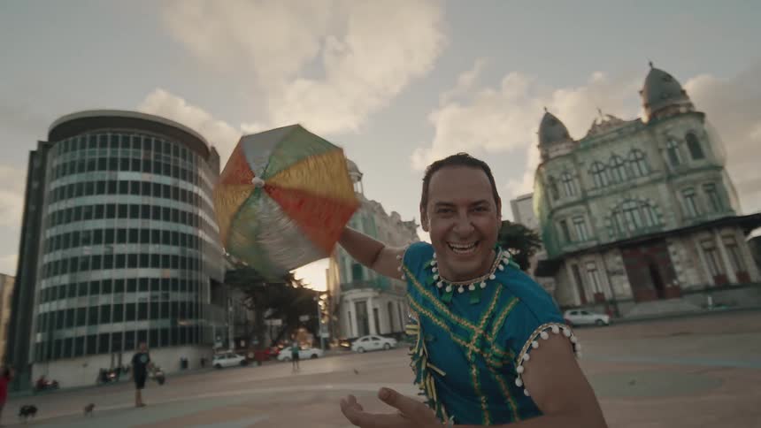 A young man wearing a blue native shirt holds a small umbrella and jumps up and down like hes dancing in the middle of the street at midday