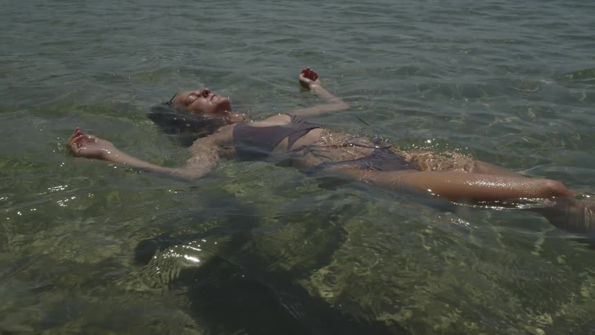A woman floats on her back in clear shallow water with her arms extended and eyes closed