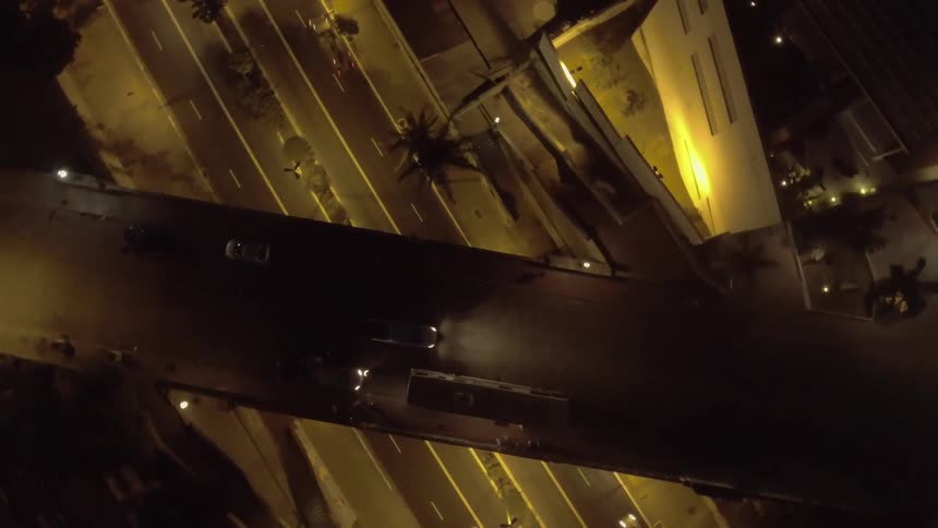 A series of cars move along a busy city road at night with an overhead bridge crossing above