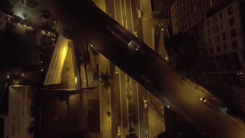 A series of cars move along a busy city road at night with an overhead bridge crossing above