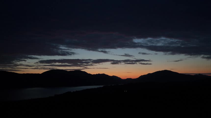 A landscape view at twilight shows dark mountain silhouettes against an orange and blue sky with a calm body of water visible below
