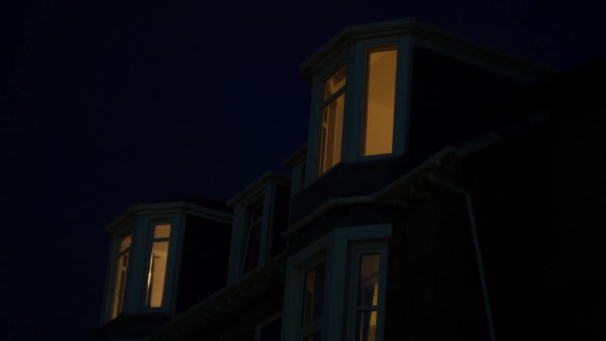 A dark house exterior is seen at night with warm light glowing from its upper windows