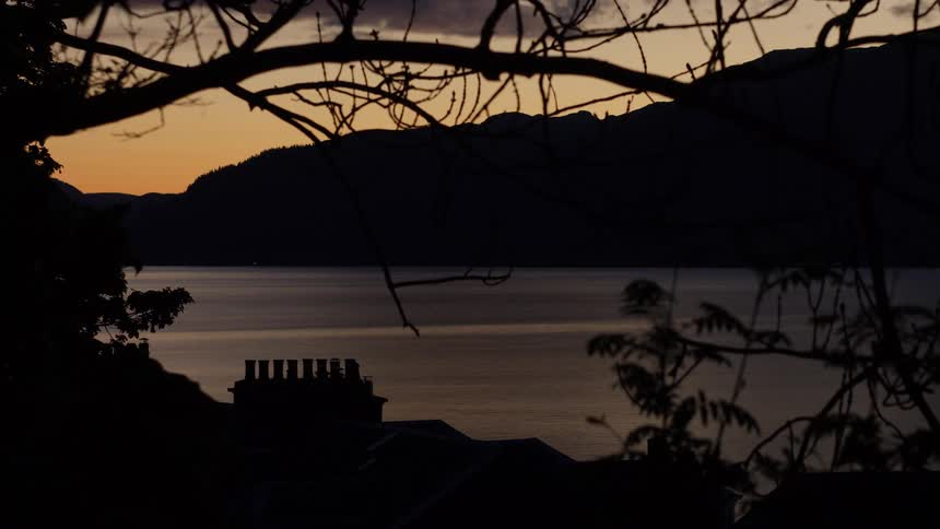 Silhouetted trees and rooftops frame a peaceful lake at sunset with distant mountains