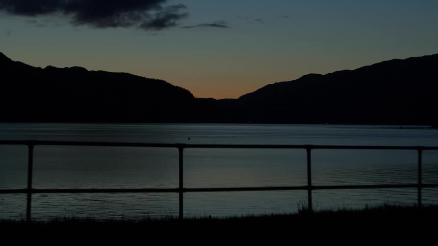 Silhouetted mountains frame a calm lake at dusk with a faint orange glow on the horizon