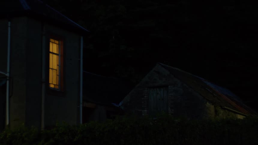House with illuminated window contrasts with dark shed next door at night