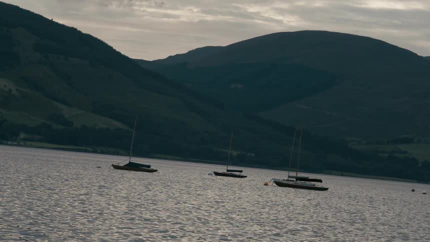 Sailboats sit calmly on a lake with mountains silhouetted in the background during dusk