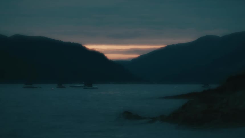 A peaceful coastal inlet at dusk with boats floating between dark mountain silhouettes
