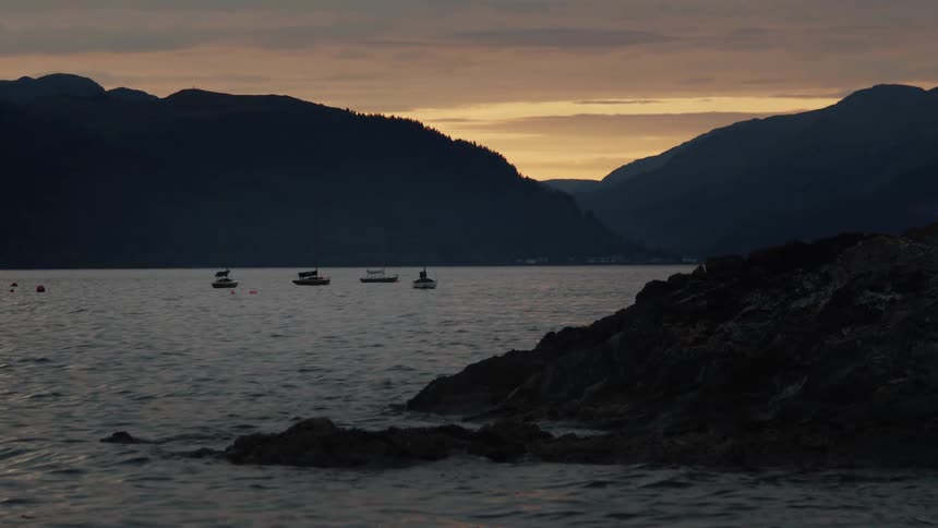 Several boats floating on a lake near distant dark mountains under a cloudy sky at dusk