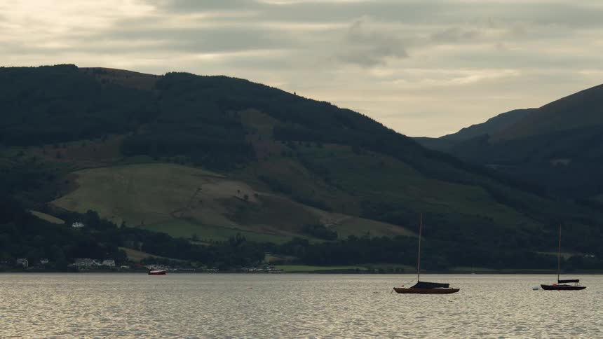 Sailboats float calmly on a lake surrounded by large green hills under an overcast sky