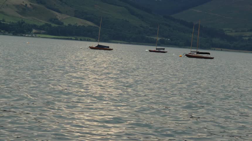 Sailboats float calmly on a lake with hills in the background under soft daylight