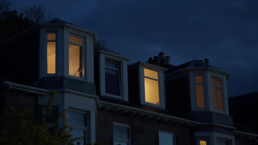 The upper part of a house is visible at night with several glowing windows and dormer roofs under a dark sky