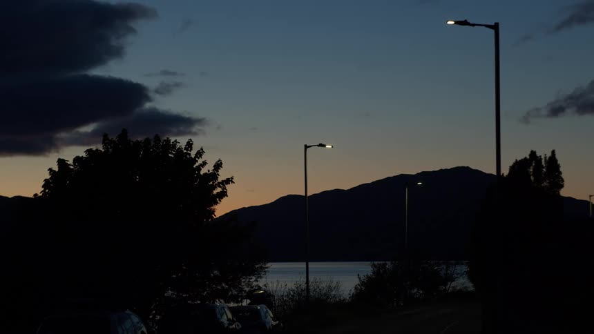 Dimly lit lakeside road at dusk with silhouetted trees and mountains
