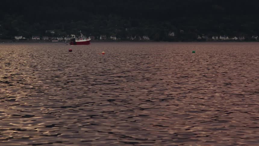 A fishing boat floats on a calm lake at sunset with buoys and distant houses lining the shoreline