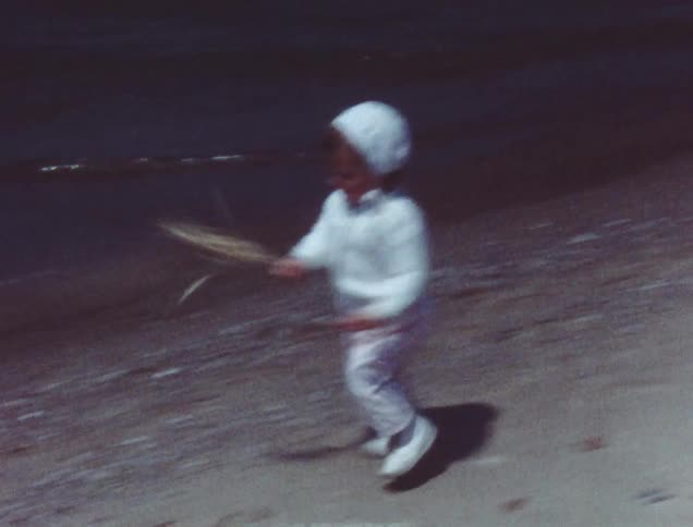 A toddler walks on the shoreline swinging a branch