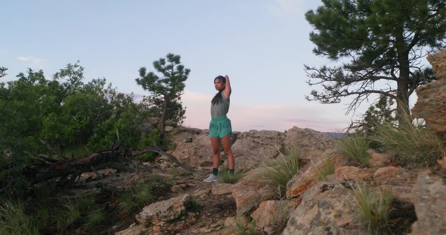 A woman stretches on a rocky trail surrounded by trees and mountainous terrain