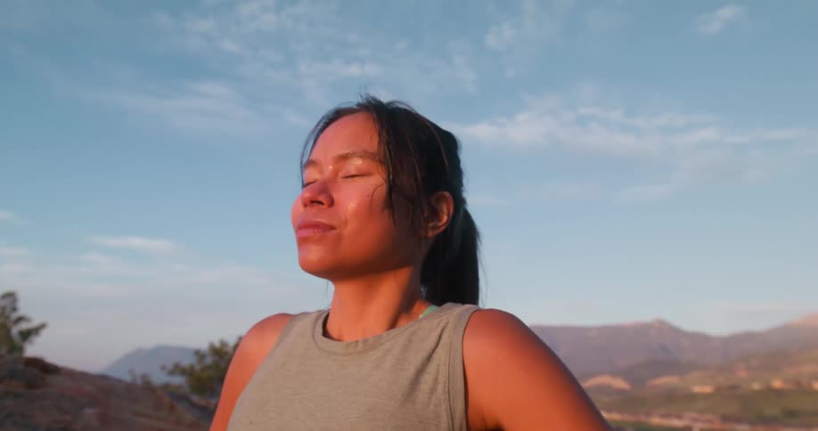 A young woman stands on top of a mountain catching her breath after a running session at midday