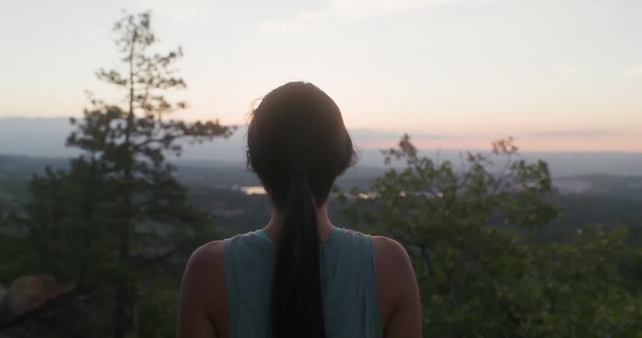 A woman stands still facing a scenic forested landscape at sunset