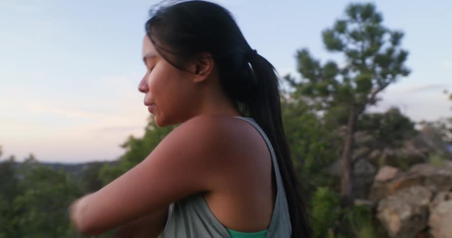 A woman stretches her arm while standing outdoors gazing into the distance at sunset