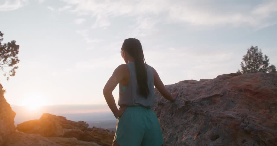 At sunset a woman wearing shorts stands beside a rock resting her hands on it as she catches her breath