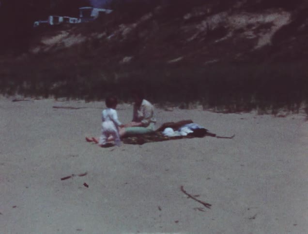 A toddler walks towards a woman sitting on the ground