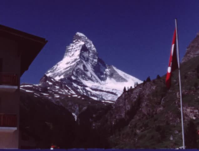 A swiss flag hangs from a flagpole with a snowy mountain peak in the distance