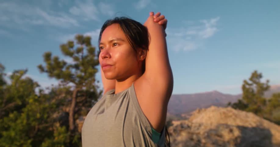 A young woman stands on a cliff in the jungle stretching her arms after a run at midday