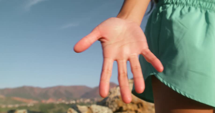 A woman stands on rocky terrain with her hands stretched out wearing green trunks at midday