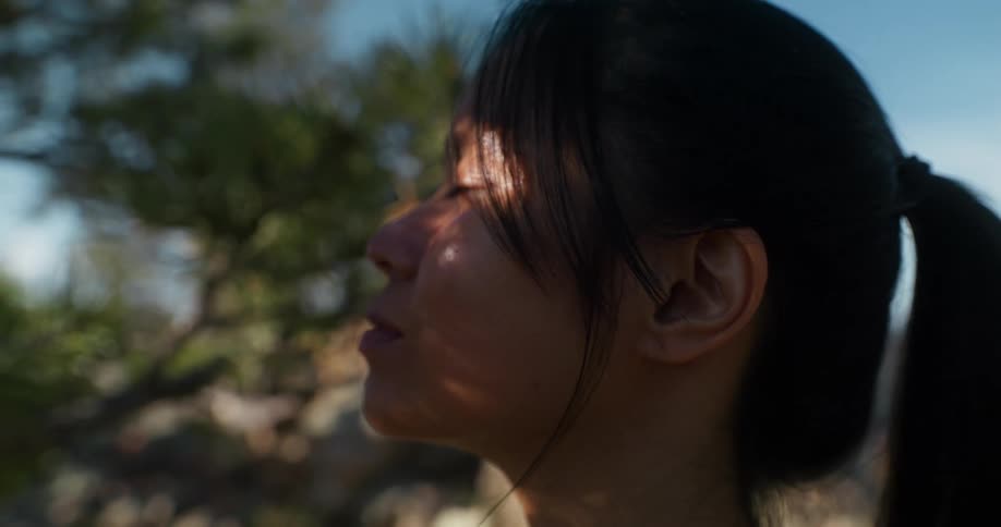 Woman with eyes closed feels the sun on her face while shadows of leaves cast over her skin then opens her eyes